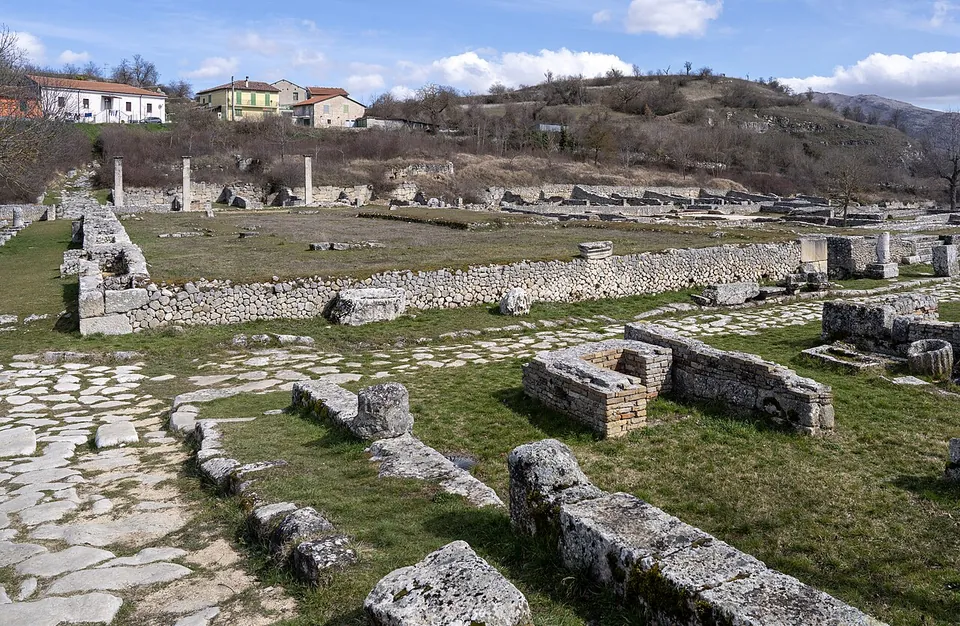A Roman street paved with large pale flagstones running between low walls of squared stone blocks, with long rows of excavated foundations and a few standing column shafts across the grass beyond, and houses on a hillside behind.
