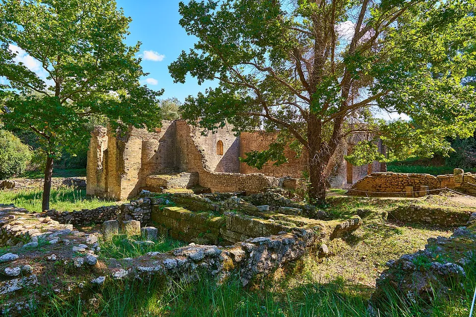 Tall ruined walls of thin red bricks with a small arched window, standing among trees above low stone foundations and grass.