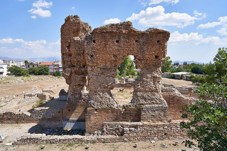 A towering ruin of red brick and rubble, its upper part pierced by an opening and arches, rising from low excavated walls under a blue sky with the modern town behind.