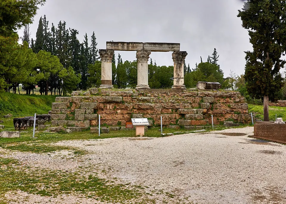 Three tall Corinthian columns carrying a stretch of beam, standing on a high podium of squared stone above a gravel square, with cypresses behind.