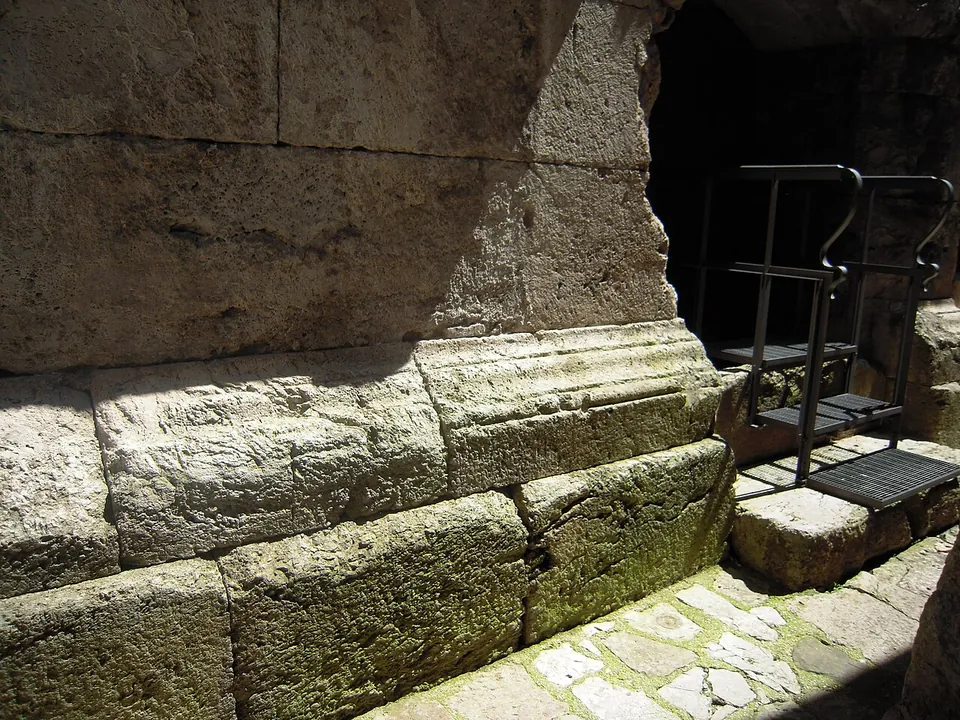 Courses of very large squared stone blocks with a projecting moulded course along their foot, a dark opening on the right with a modern iron stair, and a paved floor in front.