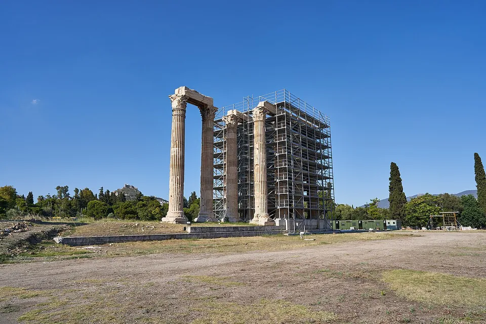The main group of columns seen across a dusty precinct, most of it wrapped in tall metal scaffolding, with two columns and a beam standing clear on the left.