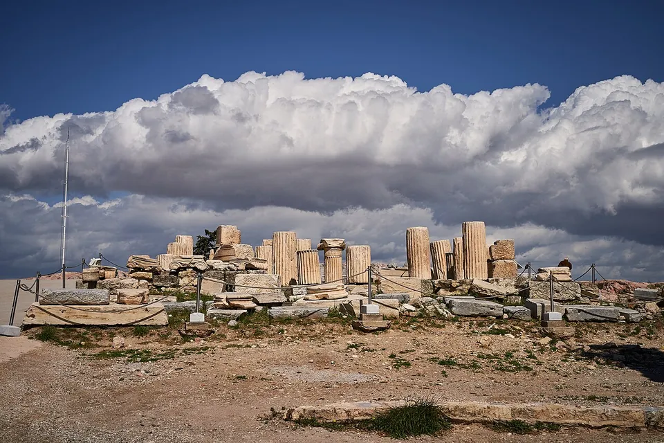 Column drums, a few short column stumps and squared marble blocks set out in rows on the bare rock behind a low rope barrier, under heavy white clouds.