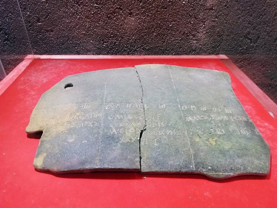 A dark green-grey plate, cracked across, incised with a grid of lines and short rows of Latin lettering, laid on red cloth.