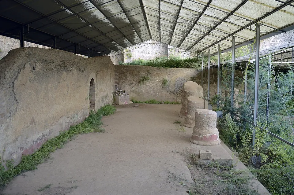 A long modern shelter of metal posts and a translucent roof covers a plastered ancient wall with a small arched opening, a row of column stumps standing on a beaten-earth floor beside it.