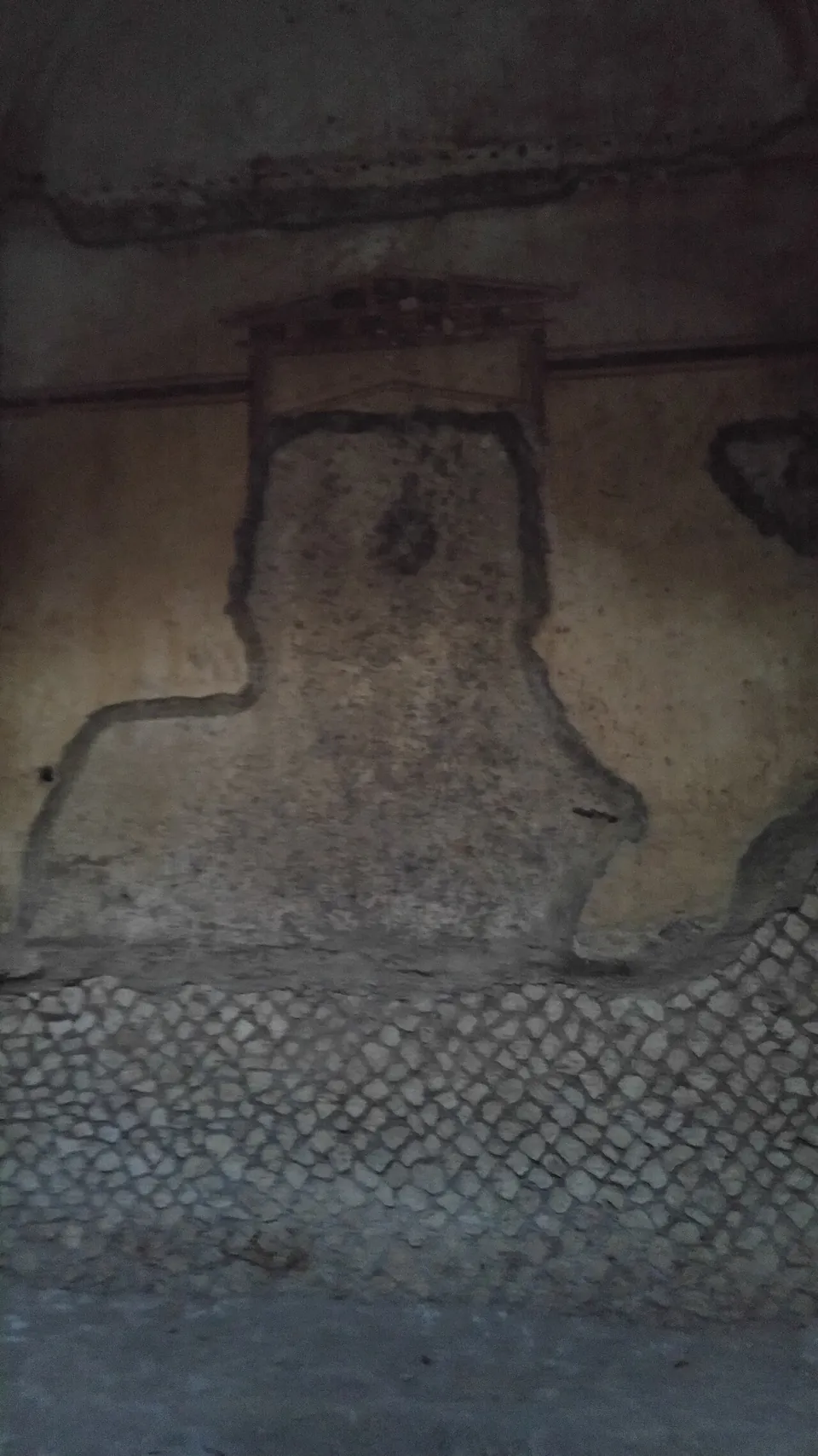 Inside the tomb, a wall with faded painted plaster in pale and dark red outlining a framed panel, and below it bare stonework laid in a net pattern.