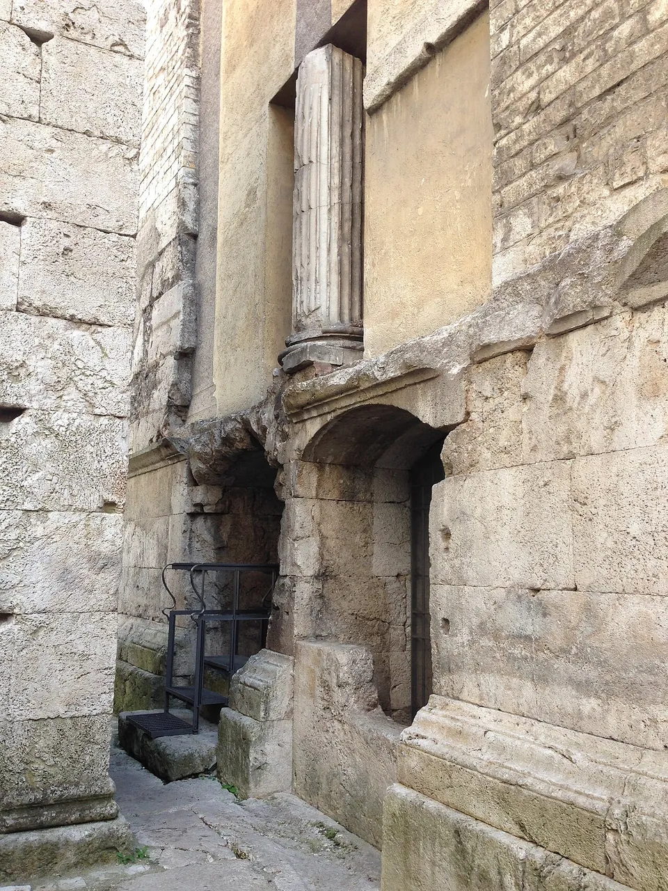 A tall wall of large squared stone blocks with the upper part of a fluted column set into it, a round-headed opening at its foot and a narrow iron stair beside it.