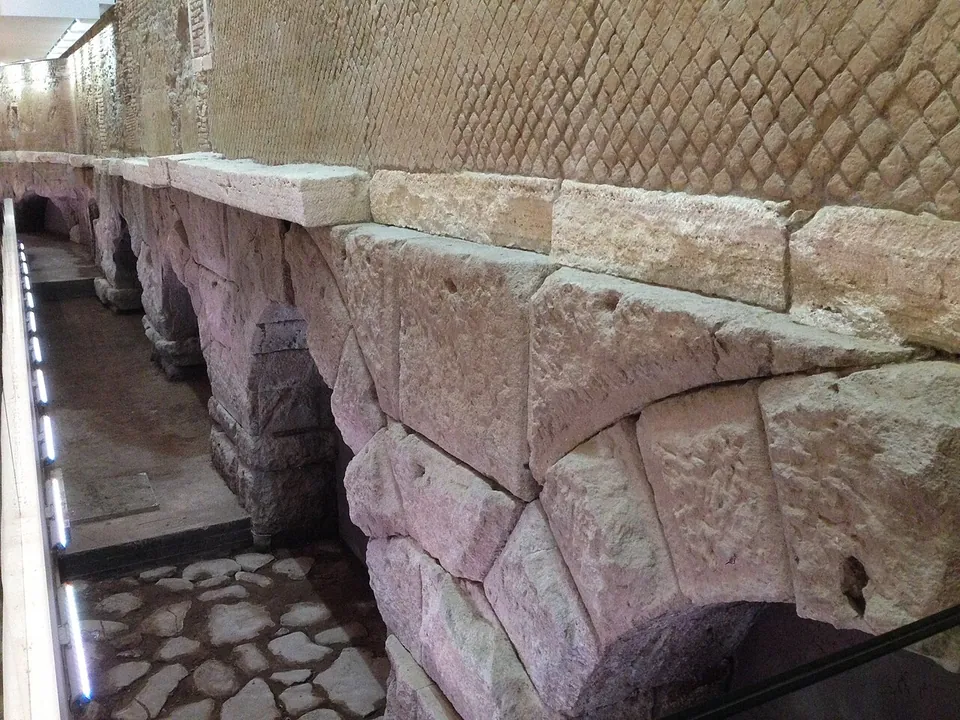 A row of low arches built of large, roughly dressed stone blocks runs along a lit gallery behind a glass edge, below a wall faced with small square stones set in a diagonal net pattern.