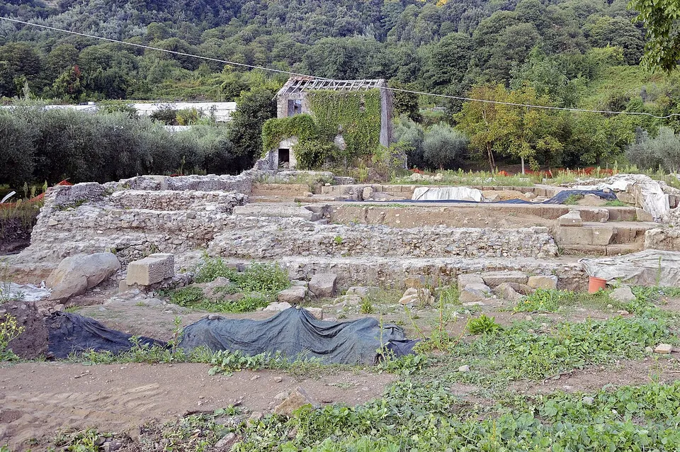 Low stepped courses of large squared stone blocks form a platform in an excavation area, with loose blocks and black protective sheeting on the ground in front and an overgrown ruined house and wooded hillside behind.