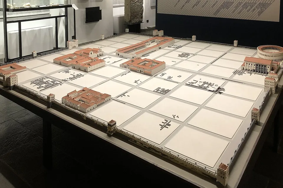 A large white table model of a rectangular walled town divided into a regular grid of blocks, with a few buildings modelled in terracotta colour, including an oval amphitheatre in one corner, displayed in a museum room.