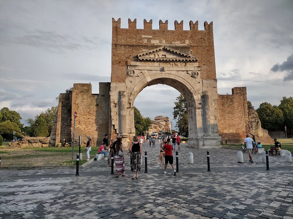 A cobbled square with bollards and people strolling towards the arch, which stands alone with short stretches of wall on either side and trees beyond, under a cloudy evening sky.