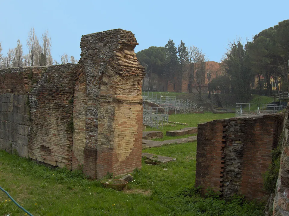 A tall pier of brick and rough stone rising from a stretch of wall at the left, with a lower brick wall at the right, on grass; trees and a brick building stand behind.