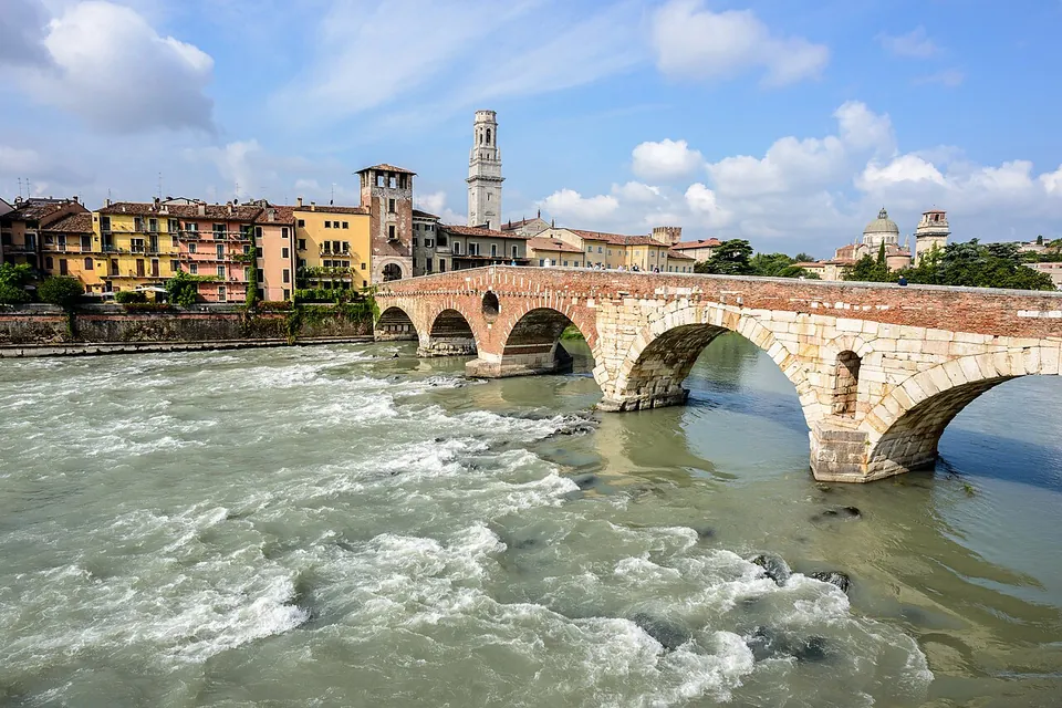 A long bridge of round arches over a fast, foaming green river. The two nearest arches are built of large pale stone blocks, with a small arched opening in the pier between them; the arches beyond are of red brick and end at a brick tower among yellow and ochre houses, with a tall bell tower behind.