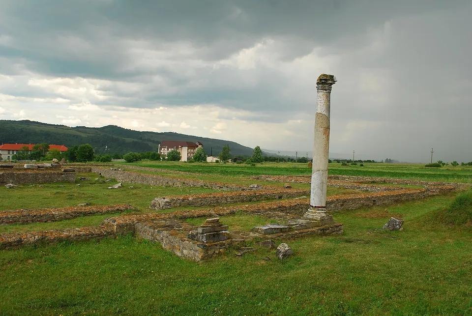 A single tall column standing among low stone foundations in a wide grassy field, with village houses and green hills under a cloudy sky.