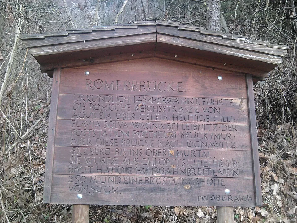 A brown wooden information board with a small roof, carved with several lines of text headed "Römerbrücke", among bare trees.