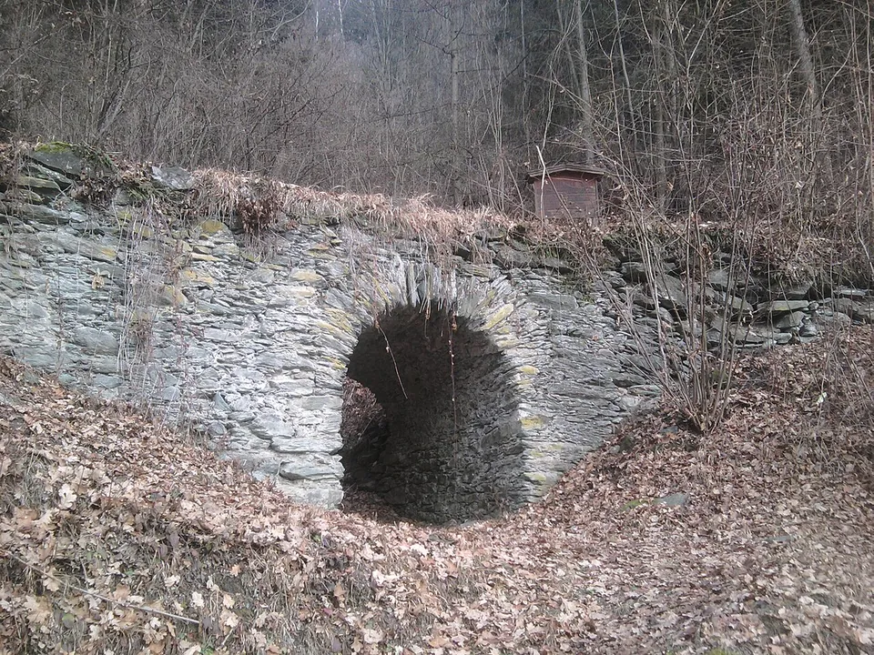 A small single-arched bridge of grey slate-like stone slabs crossing a dry, leaf-filled gully in a bare winter wood, with a small sign on top.
