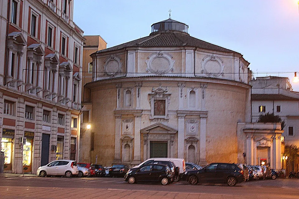 A round, domed church with a plain rendered drum and a small lantern on top, set at the end of a row of tall apartment buildings, with cars parked along the street in front at dusk.