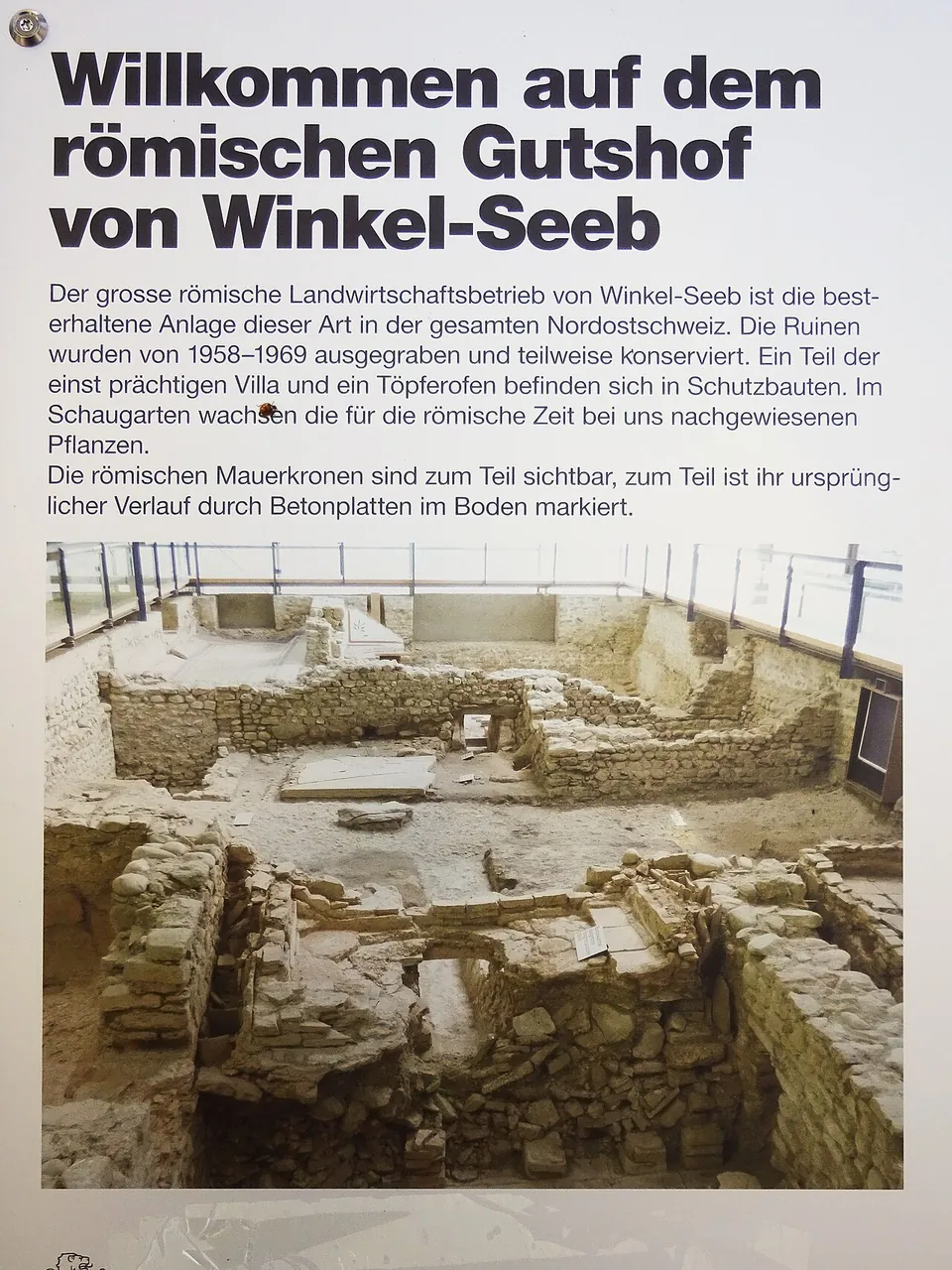 An information board headed "Willkommen auf dem römischen Gutshof von Winkel-Seeb" with four lines of German text above a photograph of excavated stone walls under a roof.