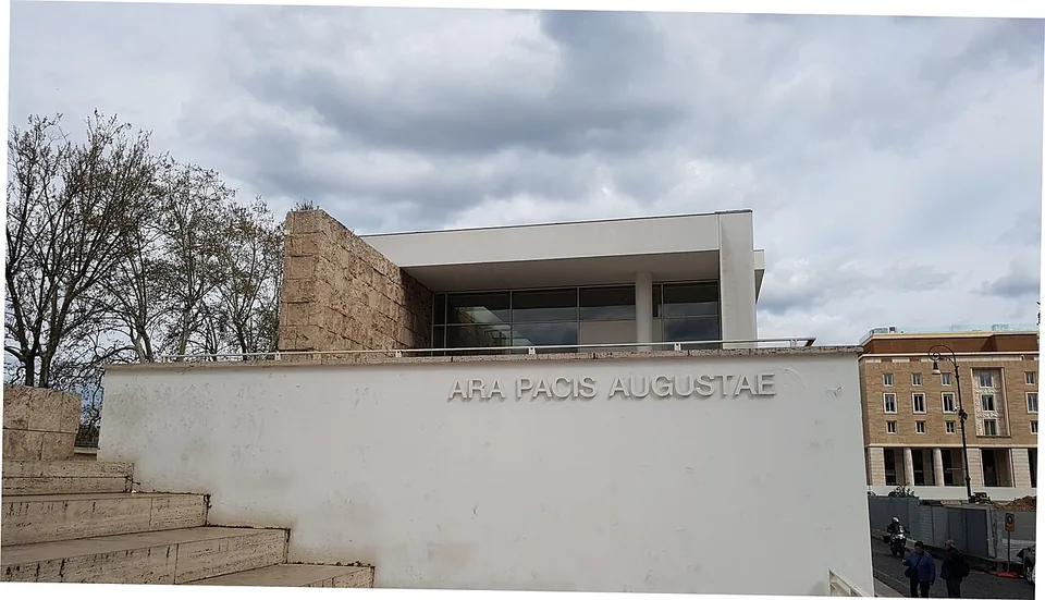 A low white wall lettered ARA PACIS AUGUSTAE in front of a modern pavilion of pale stone and glass, with steps at the left and bare trees under a cloudy sky.