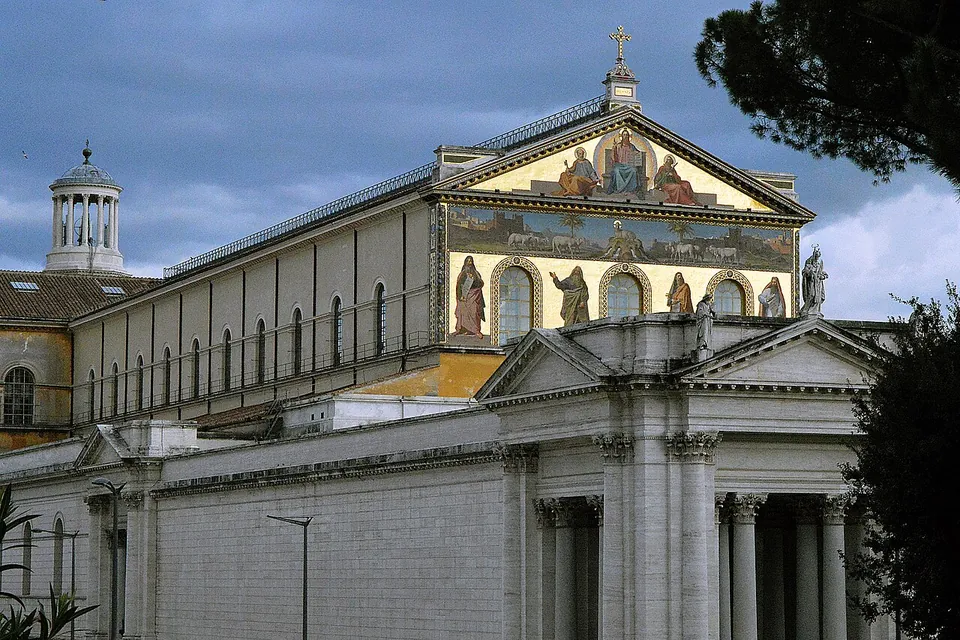 The basilica from outside under a grey sky, a long pale side wall with a row of arched windows rising to a gabled front covered in gold-ground mosaic figures, with a columned portico in the foreground and a small domed tower to the left.