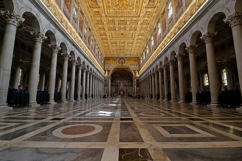 A vast nave with a patterned marble floor between long rows of tall grey columns carrying arches, a gilded coffered ceiling overhead and the triumphal arch and apse at the far end.