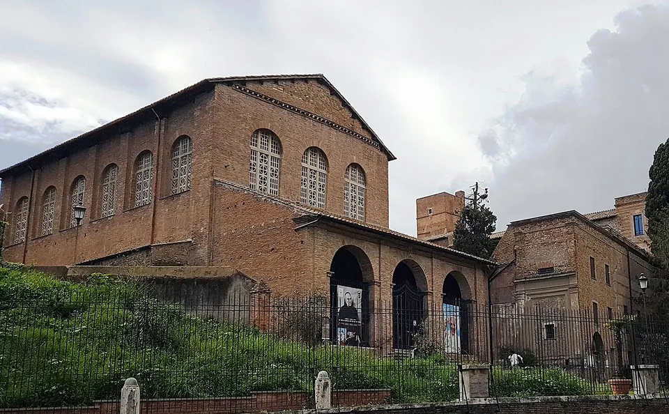 A plain brick church with a gabled roof, arched upper windows and an arcaded porch, standing above a grassy bank behind railings, with a tower and convent buildings to the right.