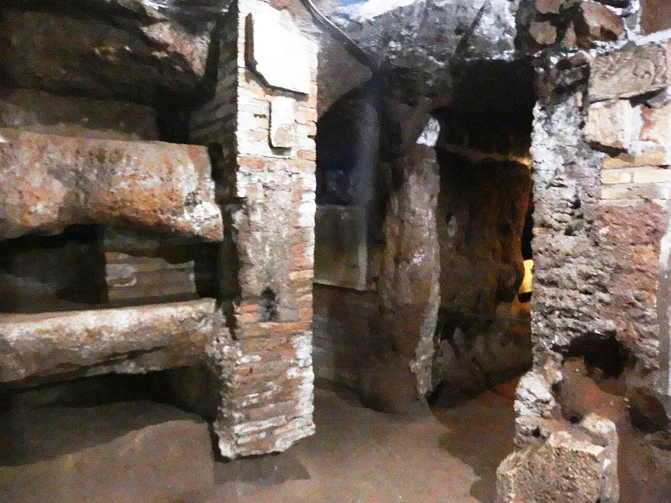 An underground passage cut in brown rock, its walls lined with long burial niches and propped by pillars of red brick, opening into further dark galleries.