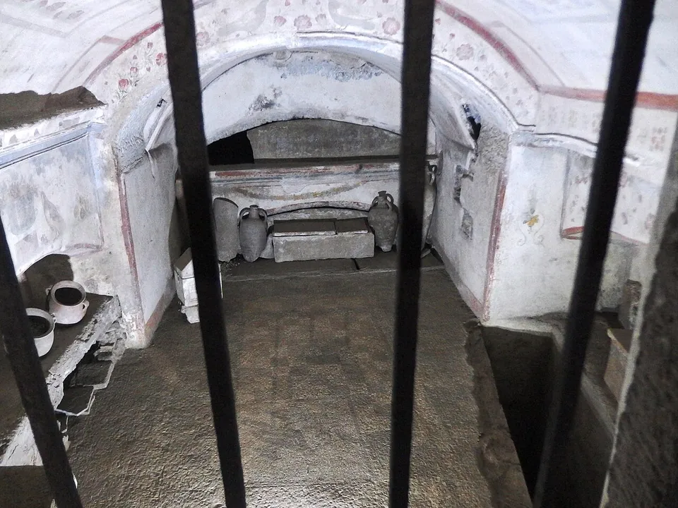 Through iron bars, a small vaulted chamber whose white walls are painted with red lines and faded motifs, with clay jars, a stone chest and arched recesses inside.