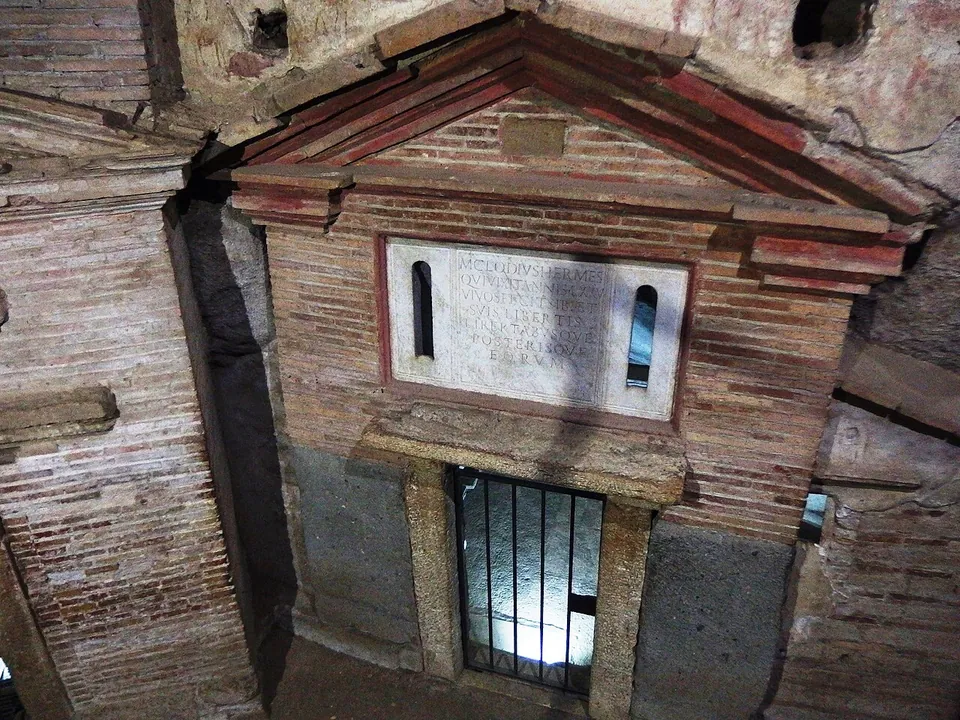 Seen from above, the front of a small brick building set against an underground rock wall, with a low brick pediment, a pale stone panel carved with several lines of Latin between two narrow windows, and a barred doorway beneath.