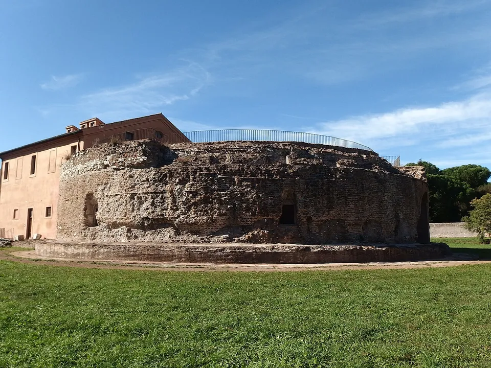 A low, round drum of rough rubble masonry with a few small openings, standing on a stone base in mown grass, with a railing along its top and a plain two-storey house rising behind it on the left.