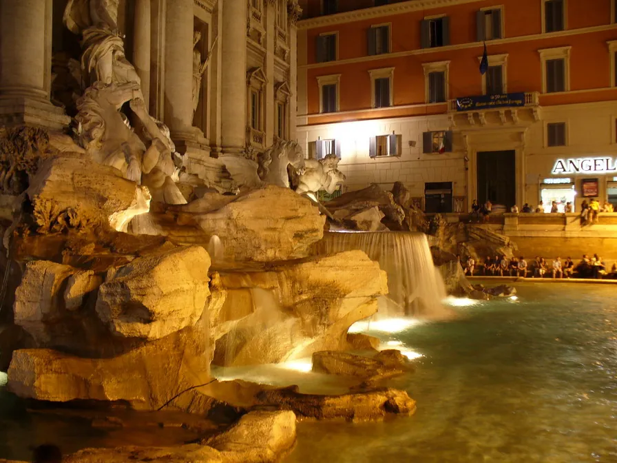 Water pouring over sculpted rocks into a wide, floodlit basin at night, with carved figures above and people sitting along the edge of the square beyond.