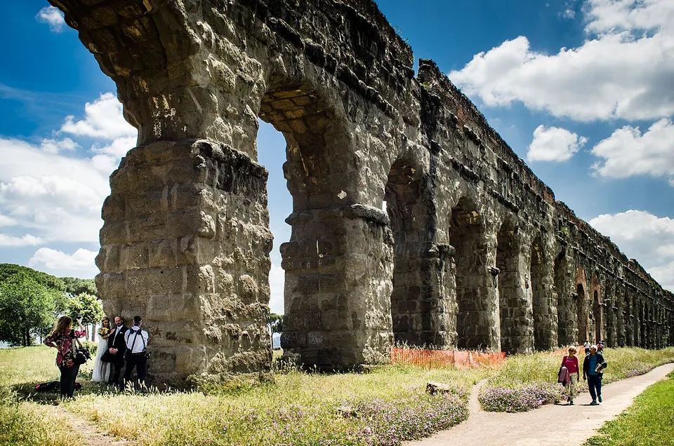 A paved path runs beside a long arcade of massive stone piers and arches, with a few people standing at the foot of the nearest pier, small against it, and flowering grass around them.