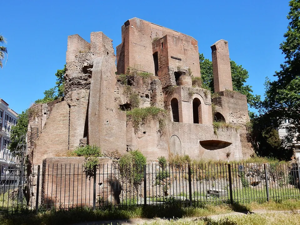 Tall ruined brick piers and walls rising on several levels, with arched niches and openings, behind a low railing among trees under a blue sky.