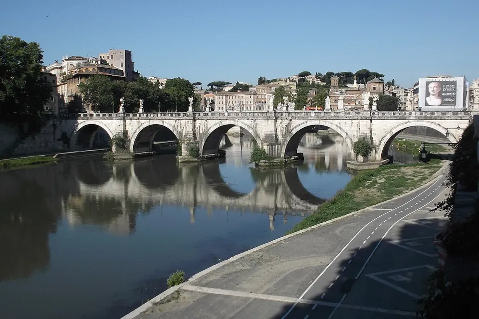 A stone bridge of five round arches across a calm river, with a row of statues along its parapet, reflected in the water, and city buildings and trees behind it.
