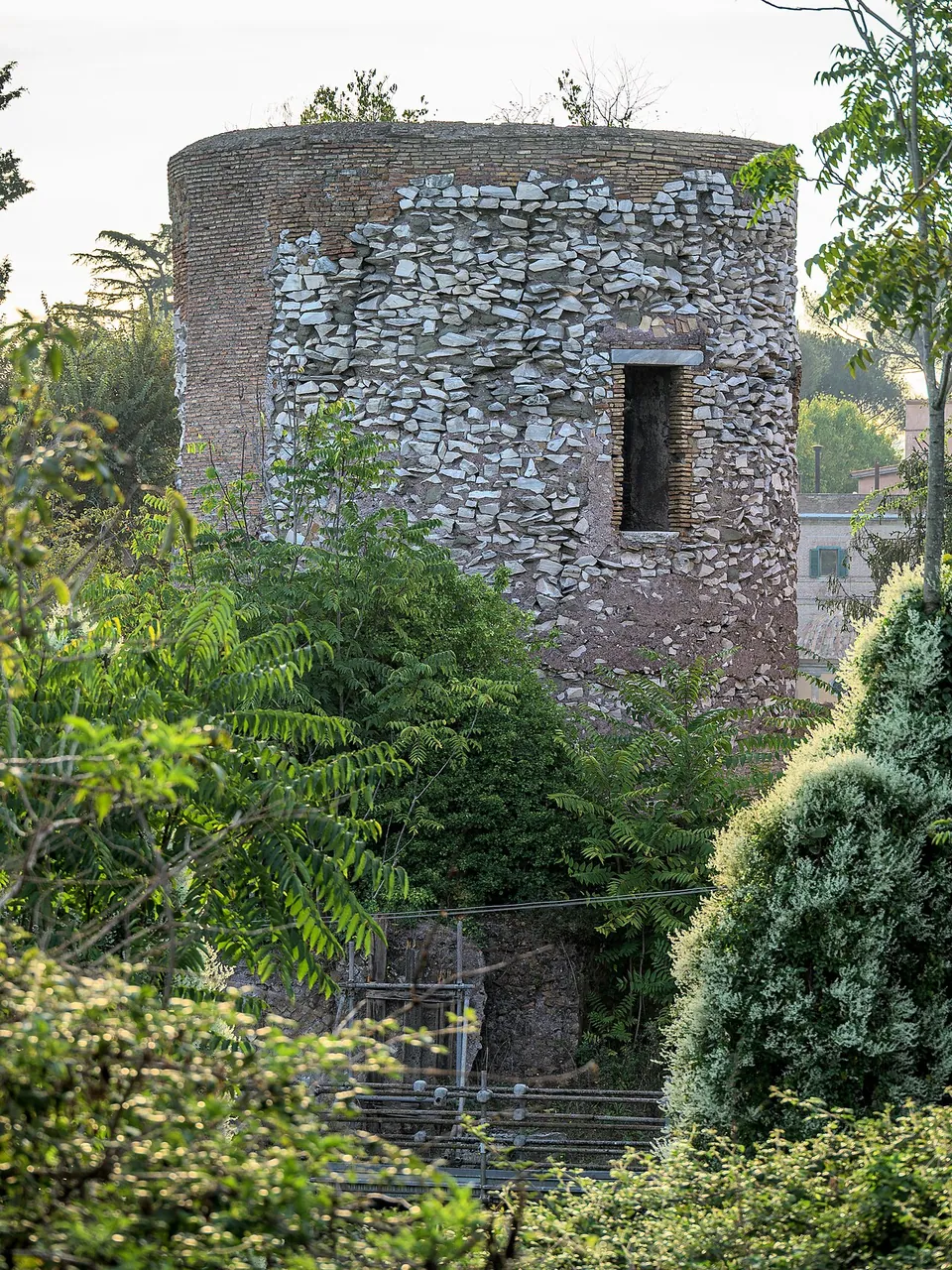 The upper cylinder of rough rubble stone, with a small rectangular window, rising above dense green shrubs and a metal railing.