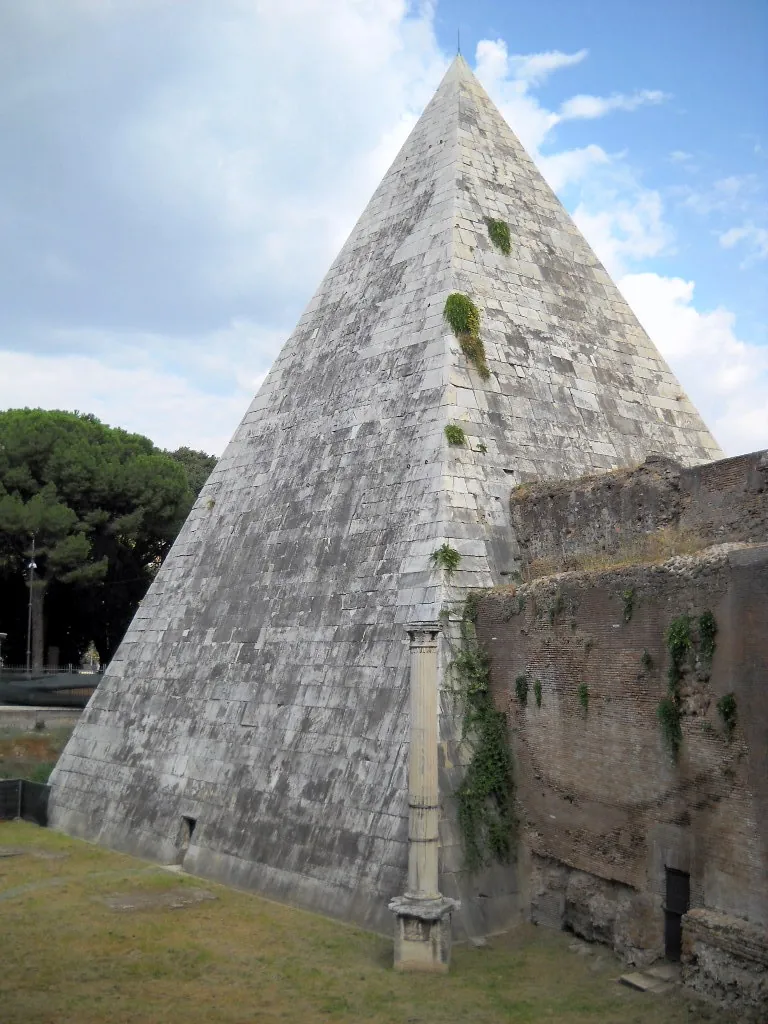 A corner of the pyramid with small plants growing from its joints, a single pale column on a pedestal standing in front of it, and the ruined brick and stone of the city wall to the right.