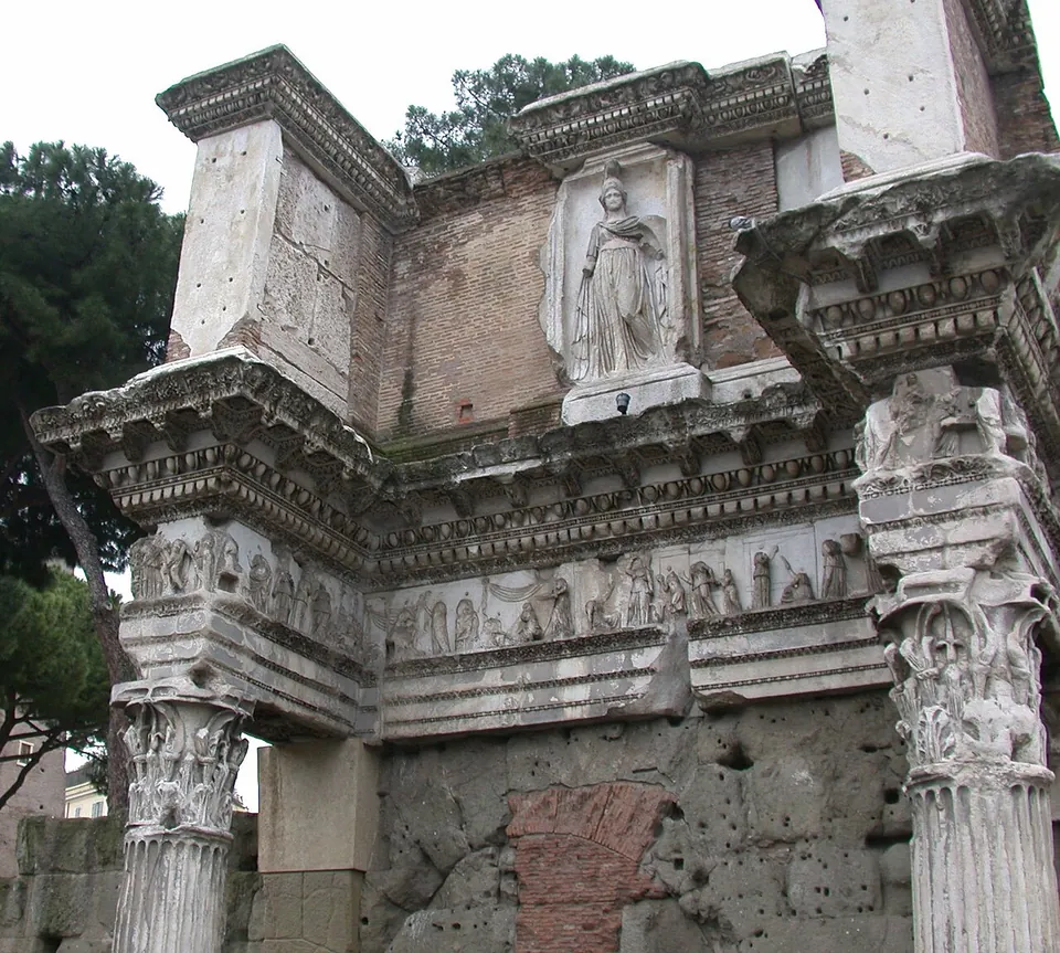 Two weathered Corinthian columns set against a wall, carrying an entablature carved with a frieze of small figures, and above it a panel with the relief of a standing robed woman wearing a helmet.