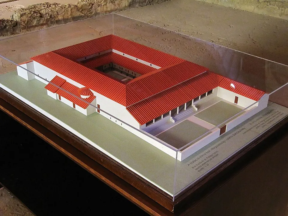 A white scale model under a glass case of a large rectangular house with red tiled roofs around an inner courtyard, with walled yards attached to one side.