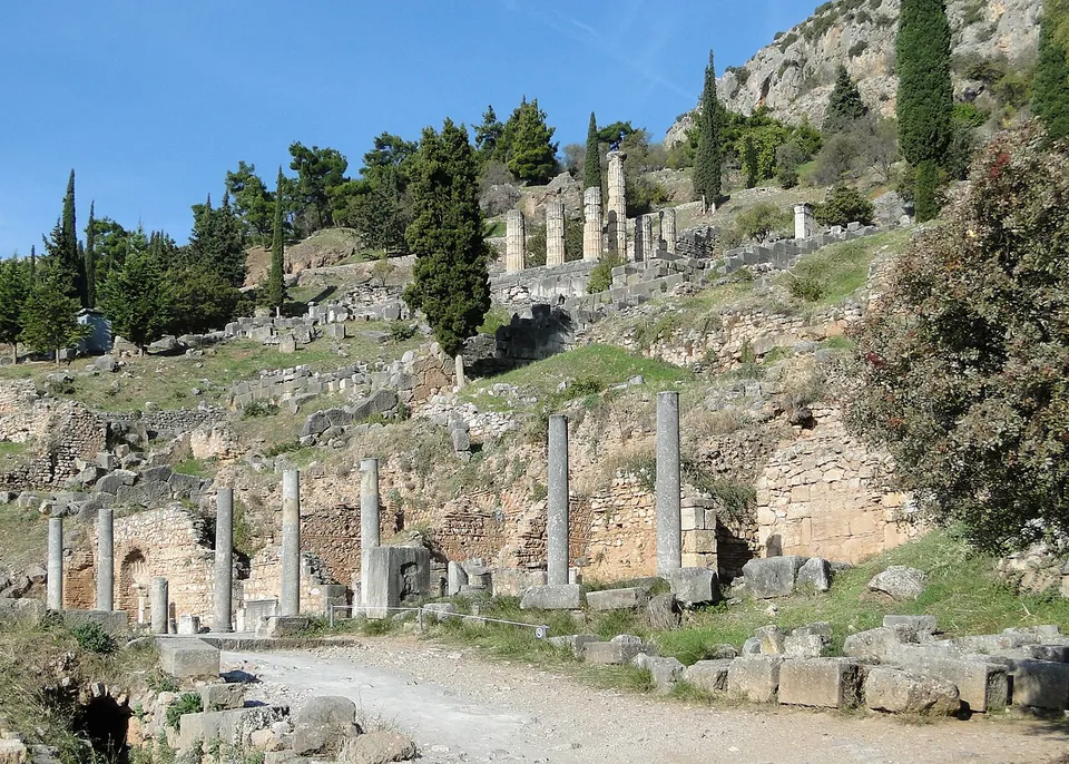 A gravel path past a row of standing column shafts and the brick and stone walls of a colonnade, with terraces of ruins, cypresses and the columns of a temple rising up the hillside behind.