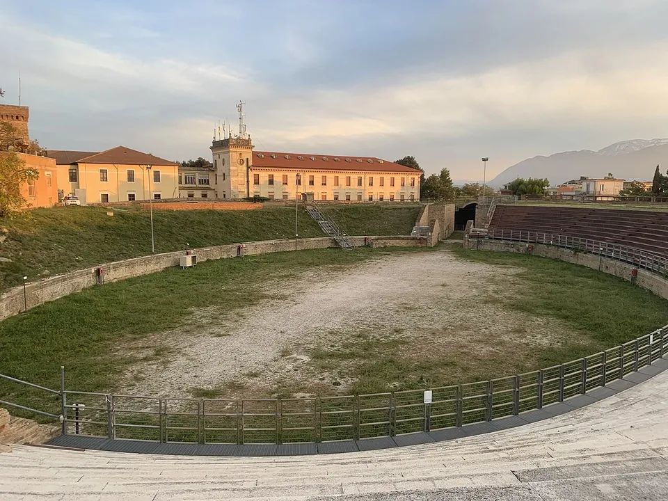 An oval arena of grass and bare earth ringed by a low wall and a metal railing, with modern stepped seating on the far side, long buildings beyond and mountains on the horizon.