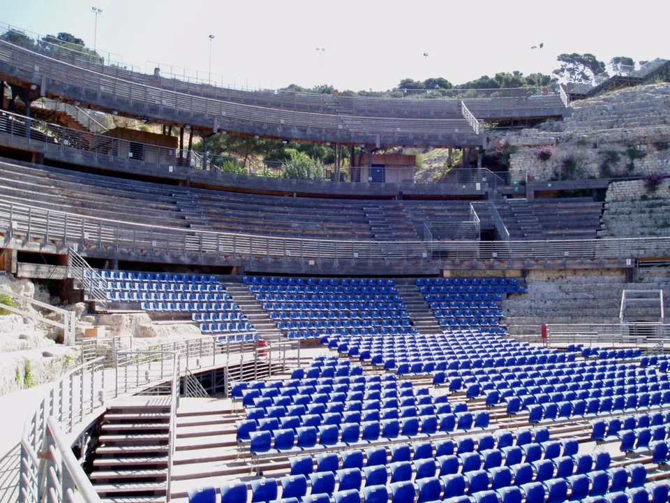 Rows of blue plastic seats on a metal and timber stand built over the ancient tiers, with walkways and a covered platform above them and the rock-cut seating visible at the edges.