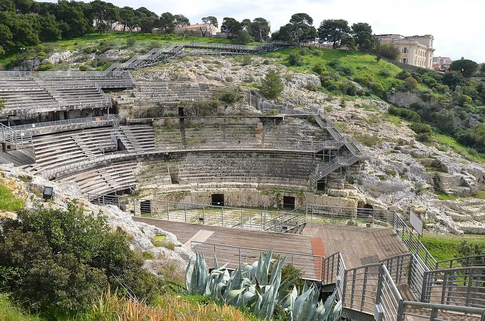 Tiers of limestone seating curving round the oval of an amphitheatre that is cut into a rocky, scrub-covered hillside, with modern metal stairways and a timber walkway running across it and trees and a large house on the ridge above.