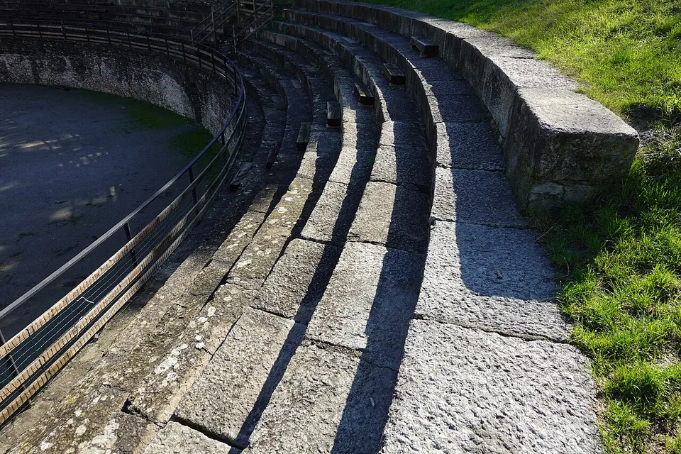 Curving tiers of large grey stone blocks seen close up in low sunlight, grass at the top edge and a metal railing along the arena wall below.