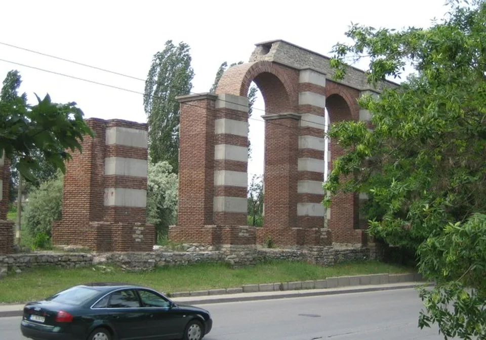 Brick piers banded with pale stone carrying two round arches, behind a low stone wall, with trees around them and a car parked on the road in front.