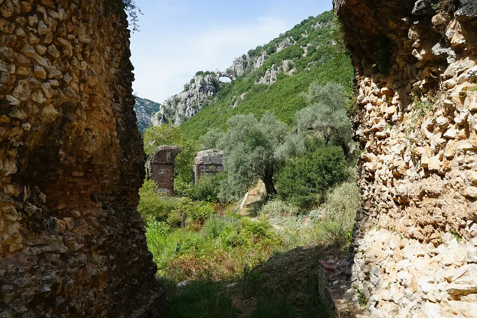 A view between two massive rubble-and-brick piers towards more piers standing among olive trees, with a steep rocky hillside behind.