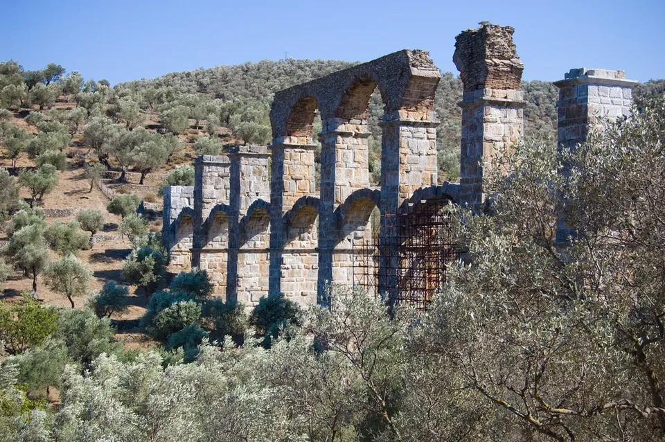 The stone arcade of the aqueduct seen through olive trees, with metal scaffolding propping one of its arches.