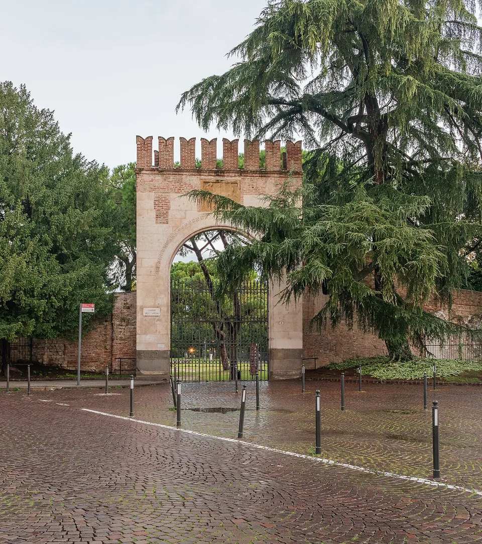 A crenellated gateway with a round arch and an iron gate, set in a wall among tall trees, seen across a wet cobbled square.