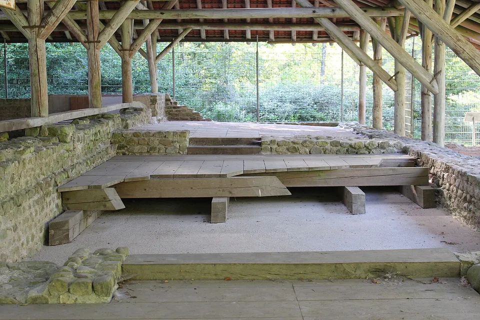 Low walls of rough stone under an open timber roof, with a raised wooden floor laid over part of the room and steps leading up behind.