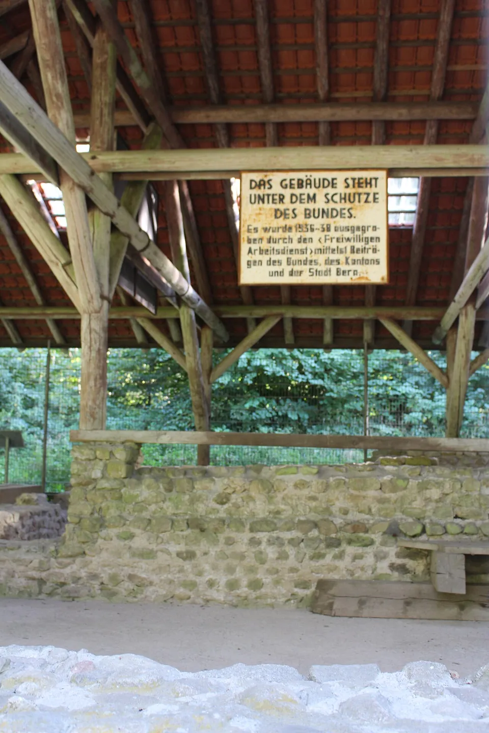 A painted sign under a timber roof above stone walls, reading in German that the building is under federal protection and was excavated in 1936-38 by the voluntary labour service with contributions from the Confederation, the canton and the city of Bern.