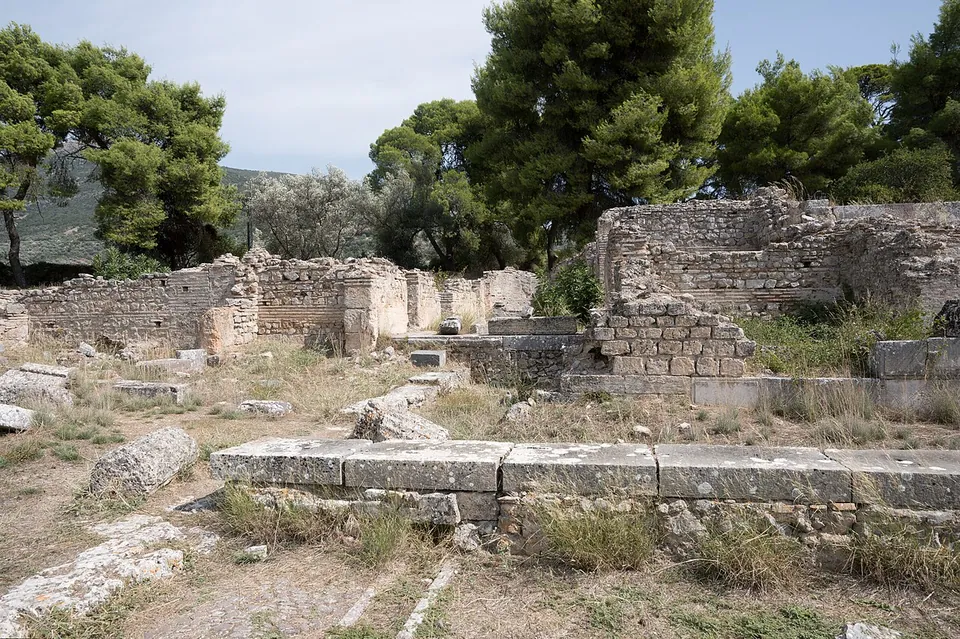Ruined walls of squared stone and rubble standing to shoulder height among pines, with a row of large stone blocks across the dry ground in front.